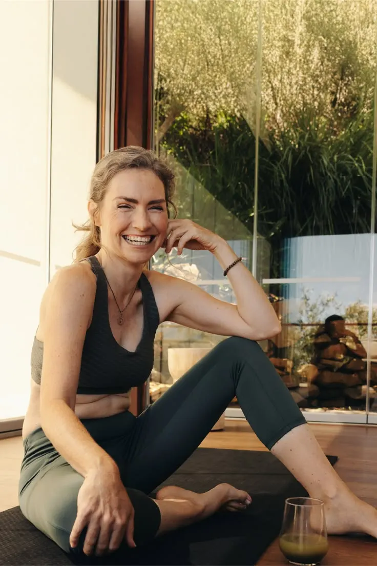 Mulher de meia-idade sorridente, sentada em tapete de ioga em ambiente com luz natural